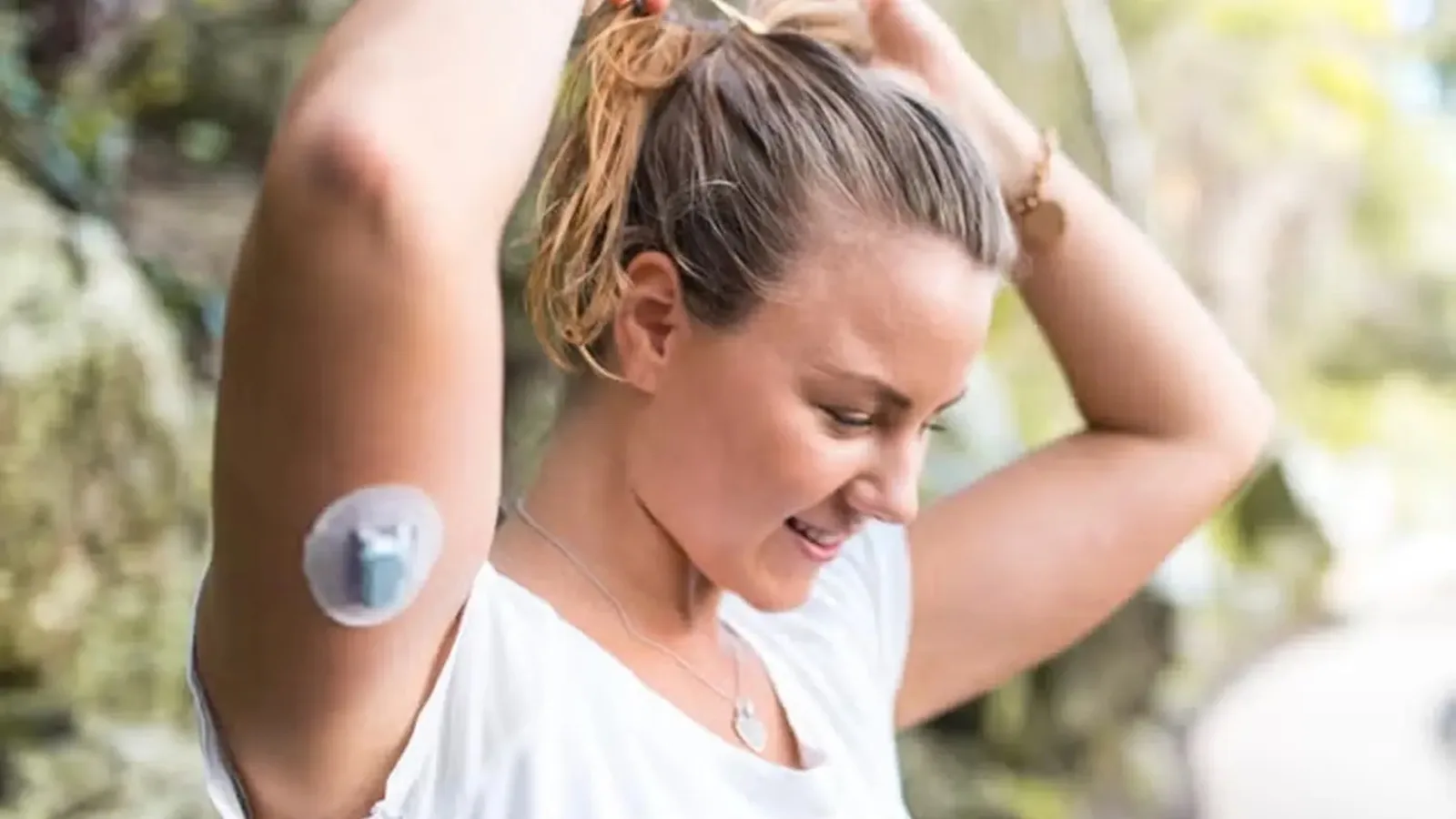 Someone outdoors tying their hair back, with a continuous glucose monitor on their upper arm